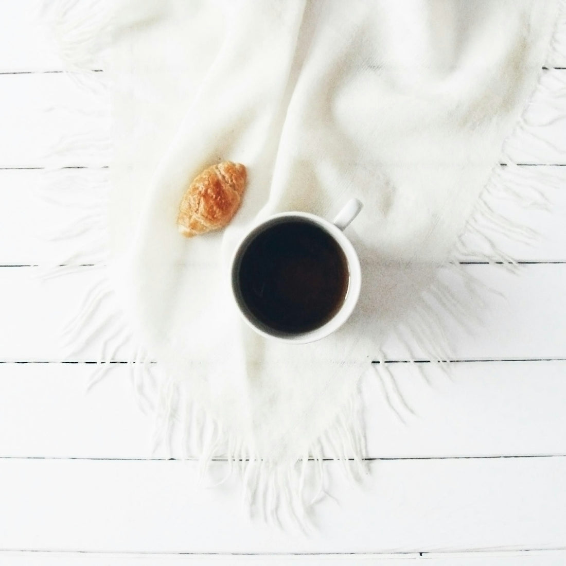 A cup of coffee and croissant on a white blanket.