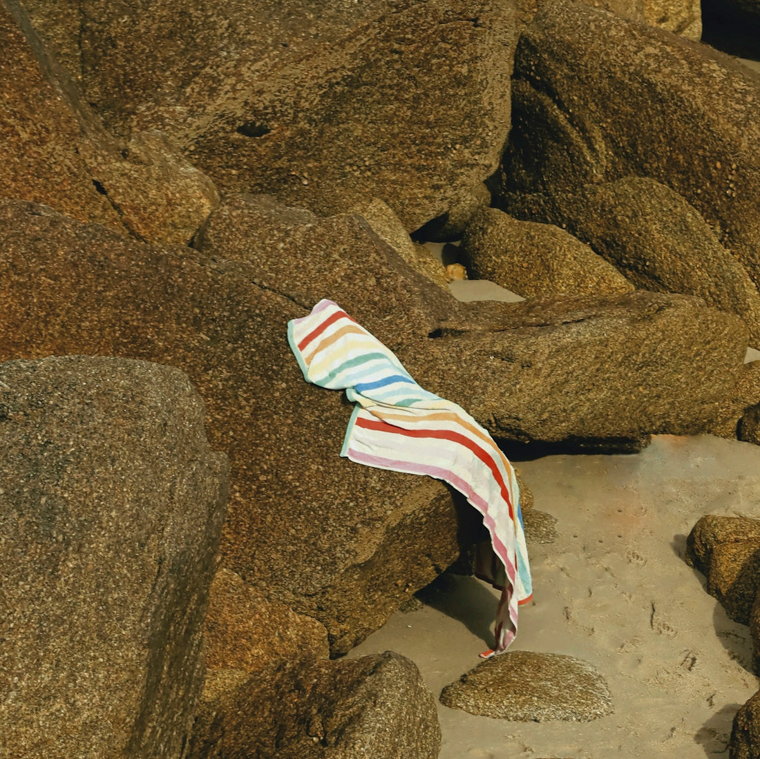 A striped beach towel draped over a large brown rock on sandy ground