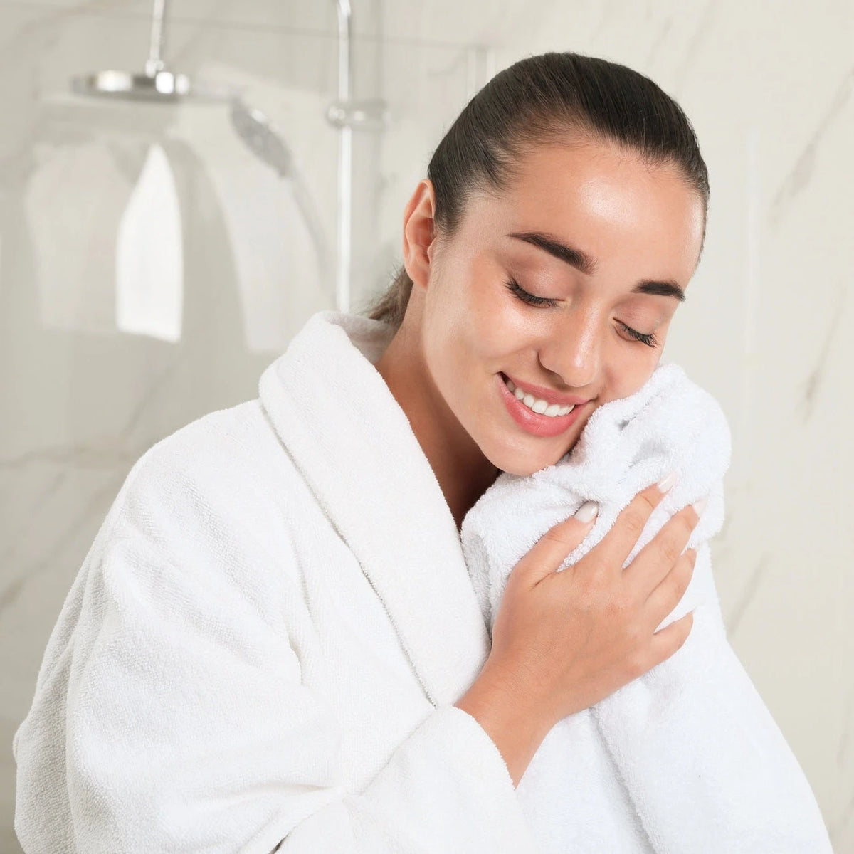 Woman in a white robe holding a towel, smiling in a bathroom setting.