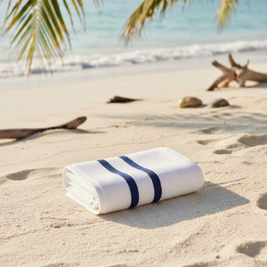 Folded white towel with blue stripes on a sandy beach with palm leaves and driftwood in the background.